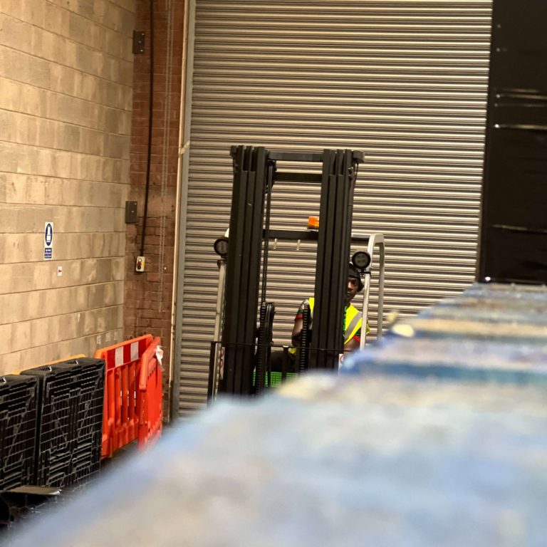 A forklift is operated in a narrow warehouse space with pallets nearby.